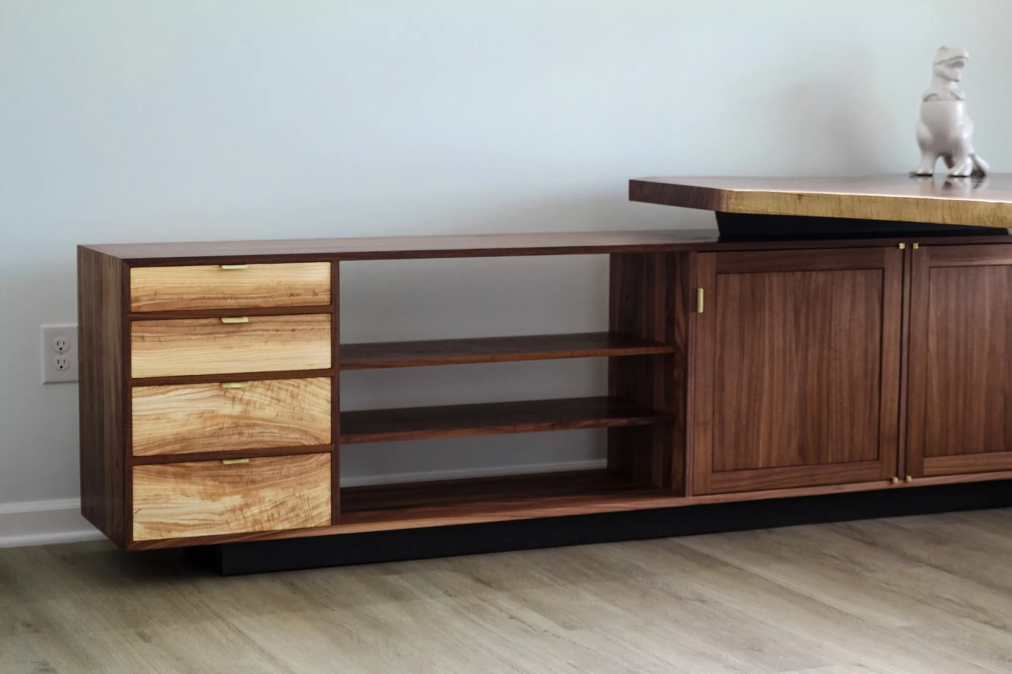 The Bottum Desk — a live-edge walnut top on a dark pedestal above a long low walnut cabinet with ambrosia maple drawer fronts, open shelves, and cabinet doors on a black base.