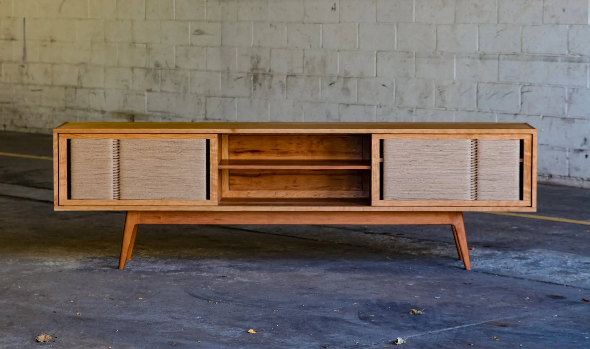 The Arnold Credenza viewed straight on, its two woven-cord doors slid apart to reveal open shelving in the center, standing on splayed tapered legs.