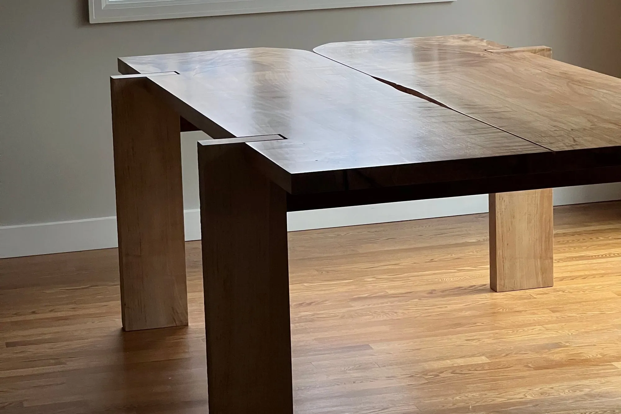 The Mueller Table in a bright room — a silver maple dining table whose top is split by a deliberate gap down the center, standing on figured silver maple legs over an oak floor.