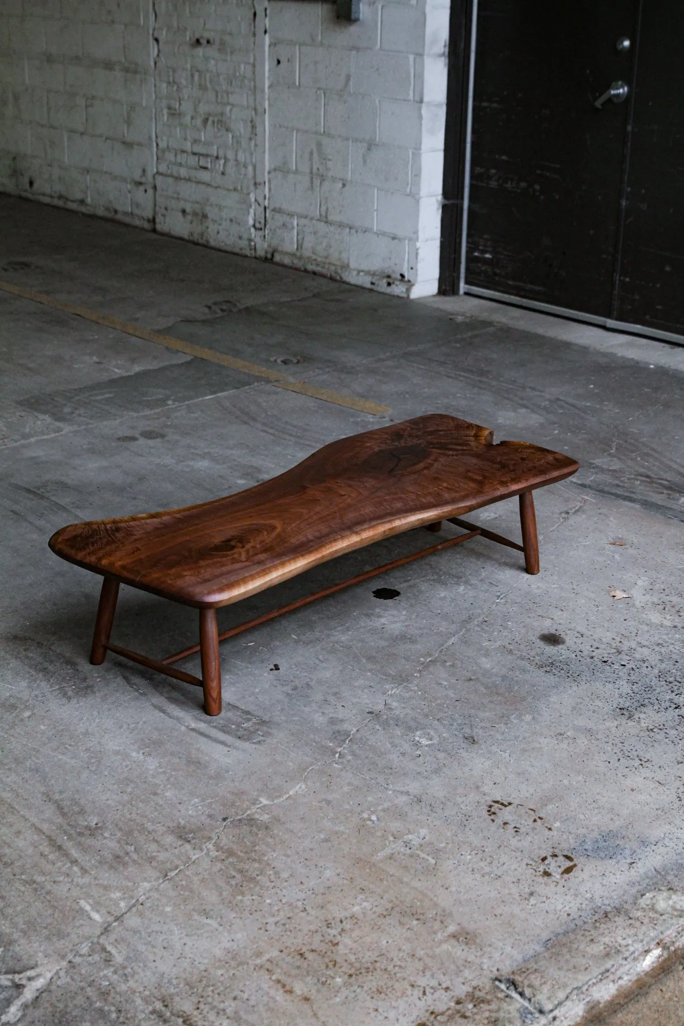 A long low walnut table with a live-edge top and turned tapered legs, standing on a concrete floor against a white brick wall.