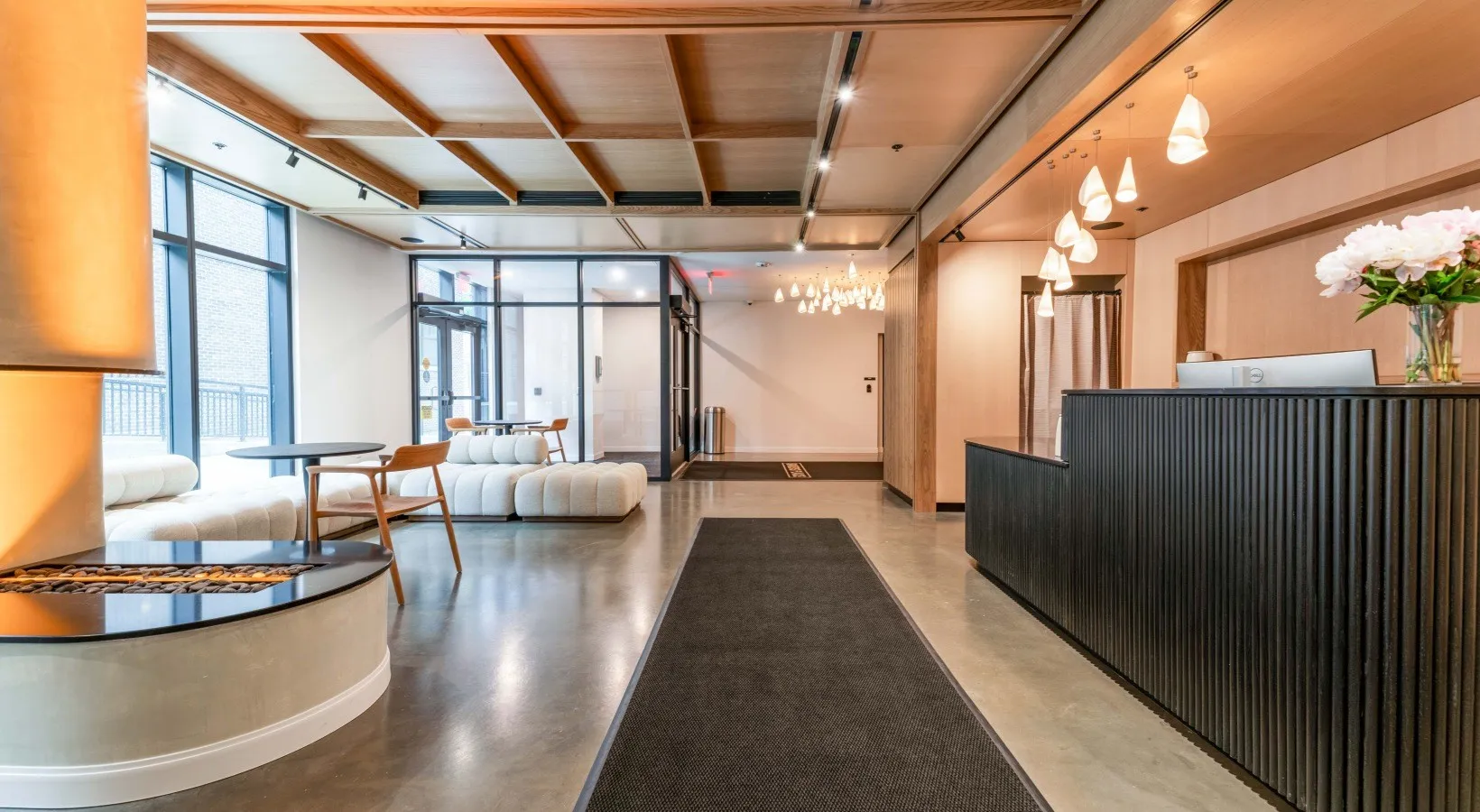 The Bakers Place lobby — a coffered wood ceiling over a polished concrete floor, with a black reeded reception desk on one side and a lounge seating area and fireplace on the other.