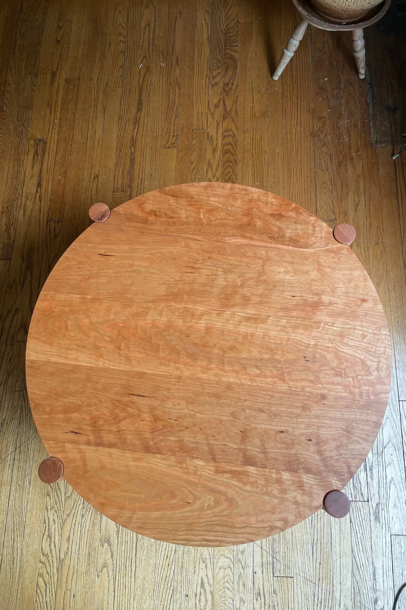 A round solid-cherry coffee table seen from directly above on a wood floor, four small round leg tenons showing through the surface near the edge.