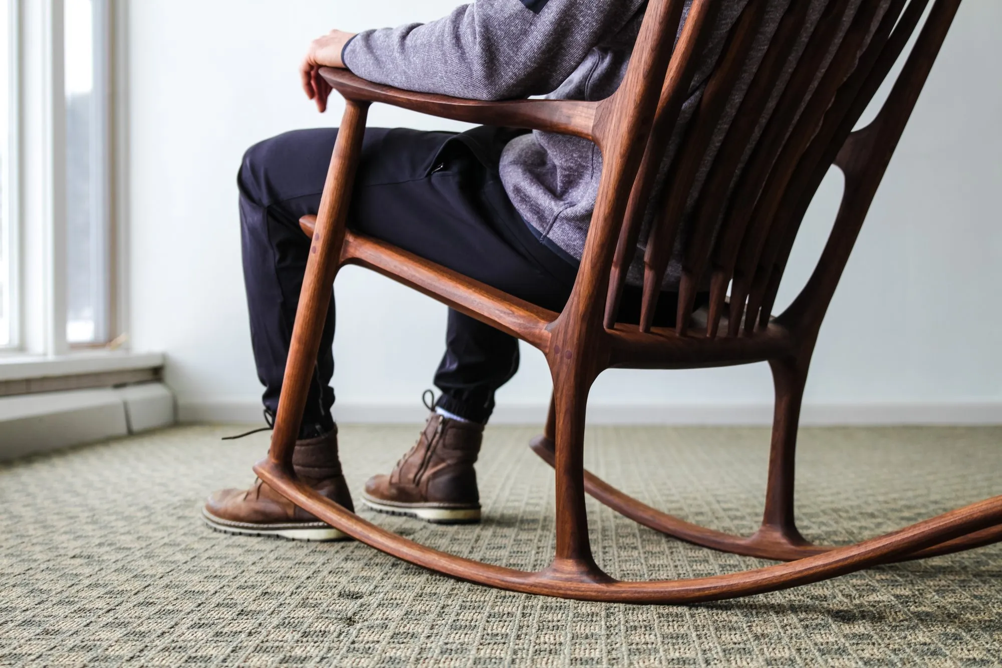 The walnut Maloof-style rocking chair in use, seen from the side as someone sits in it, showing the shaped arm, spindle back, and long curved rocker on the floor.