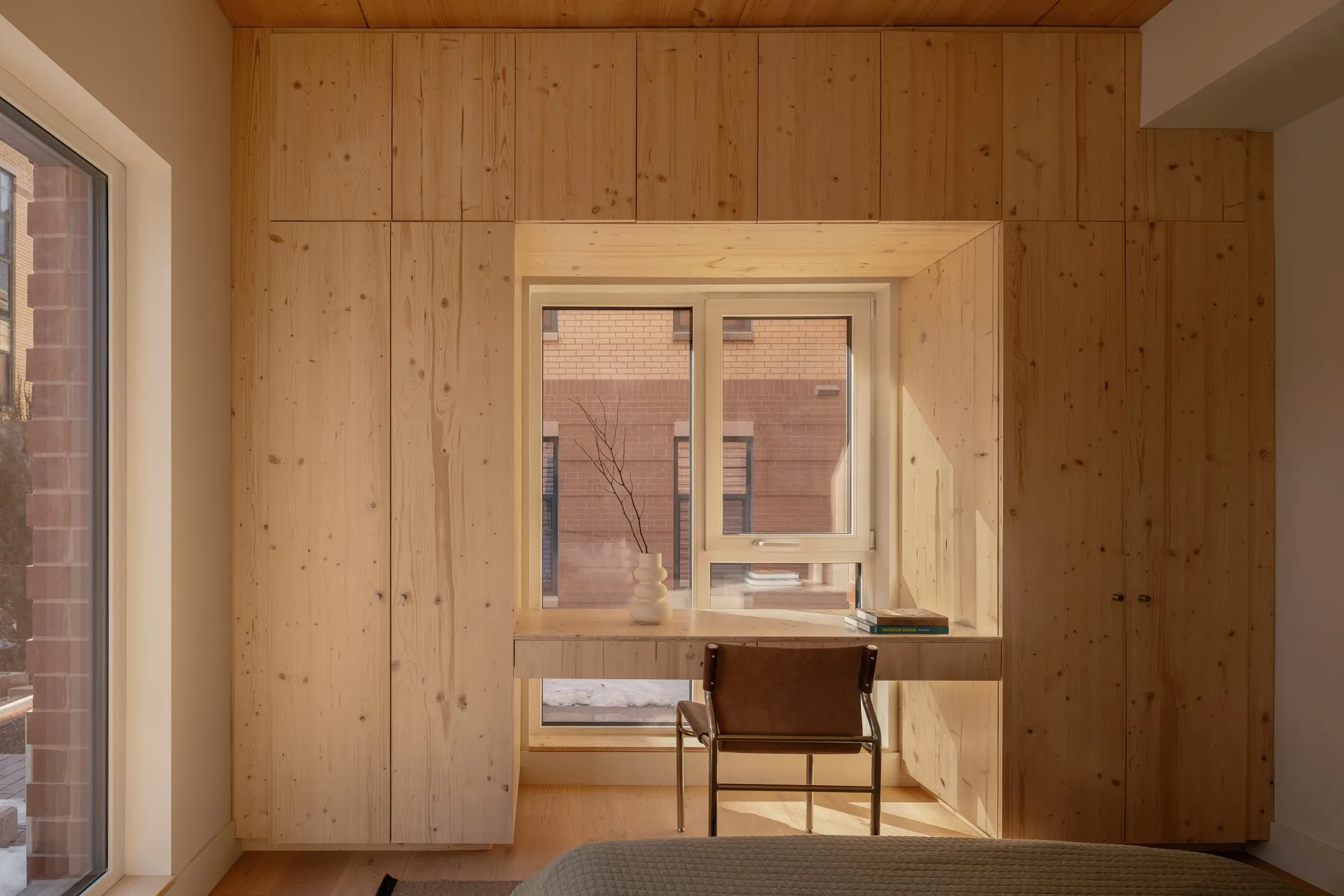A wall of built-in pine wardrobes wrapping a window, with a slim desk set into the window reveal and a leather chair.