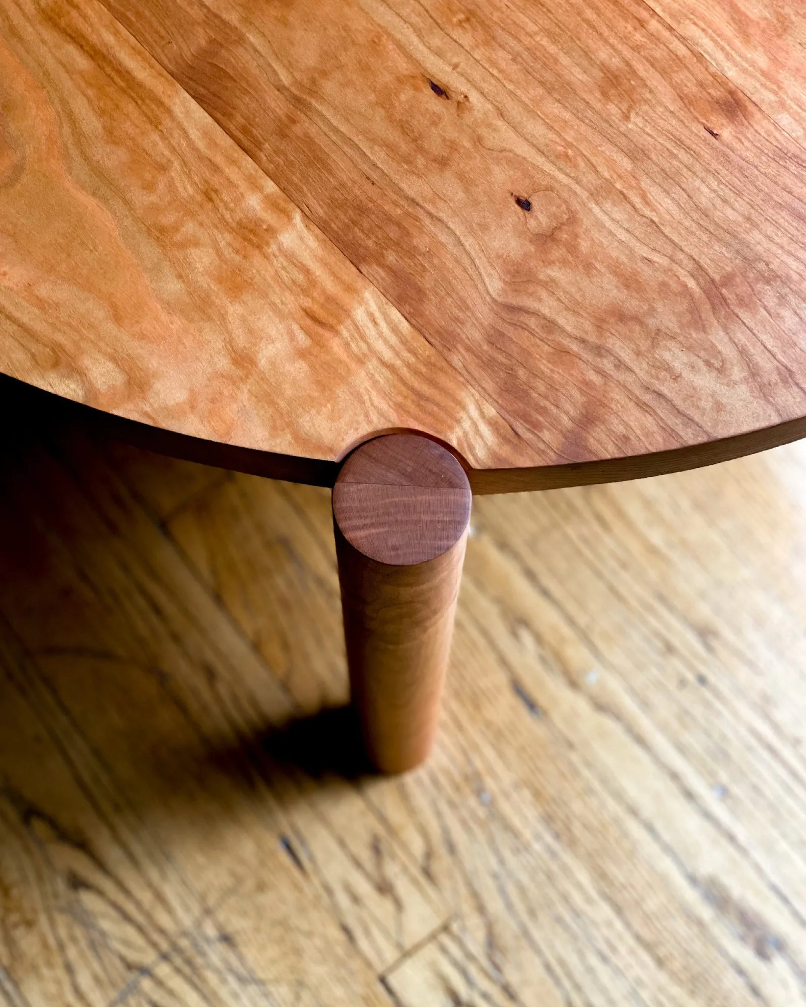 Close detail of one turned cylindrical leg meeting the underside of the round cherry top, its end capped by an exposed circular tenon flush with the surface.
