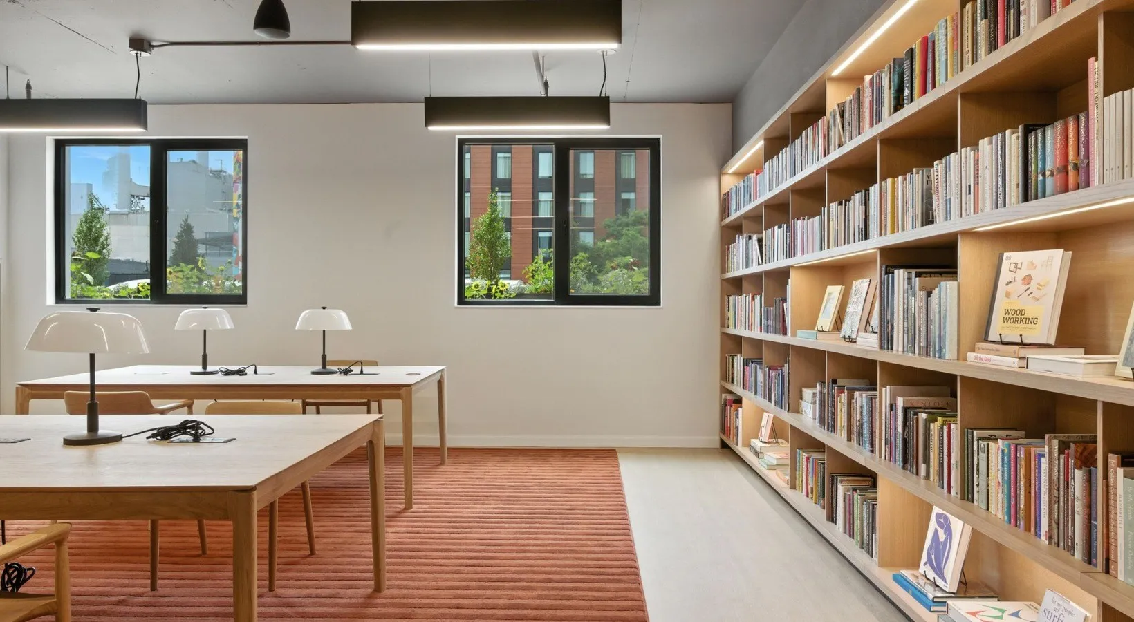 The amenity library — a full wall of built-in oak shelving beside wood worktables with task lamps, lit by windows over the neighborhood.