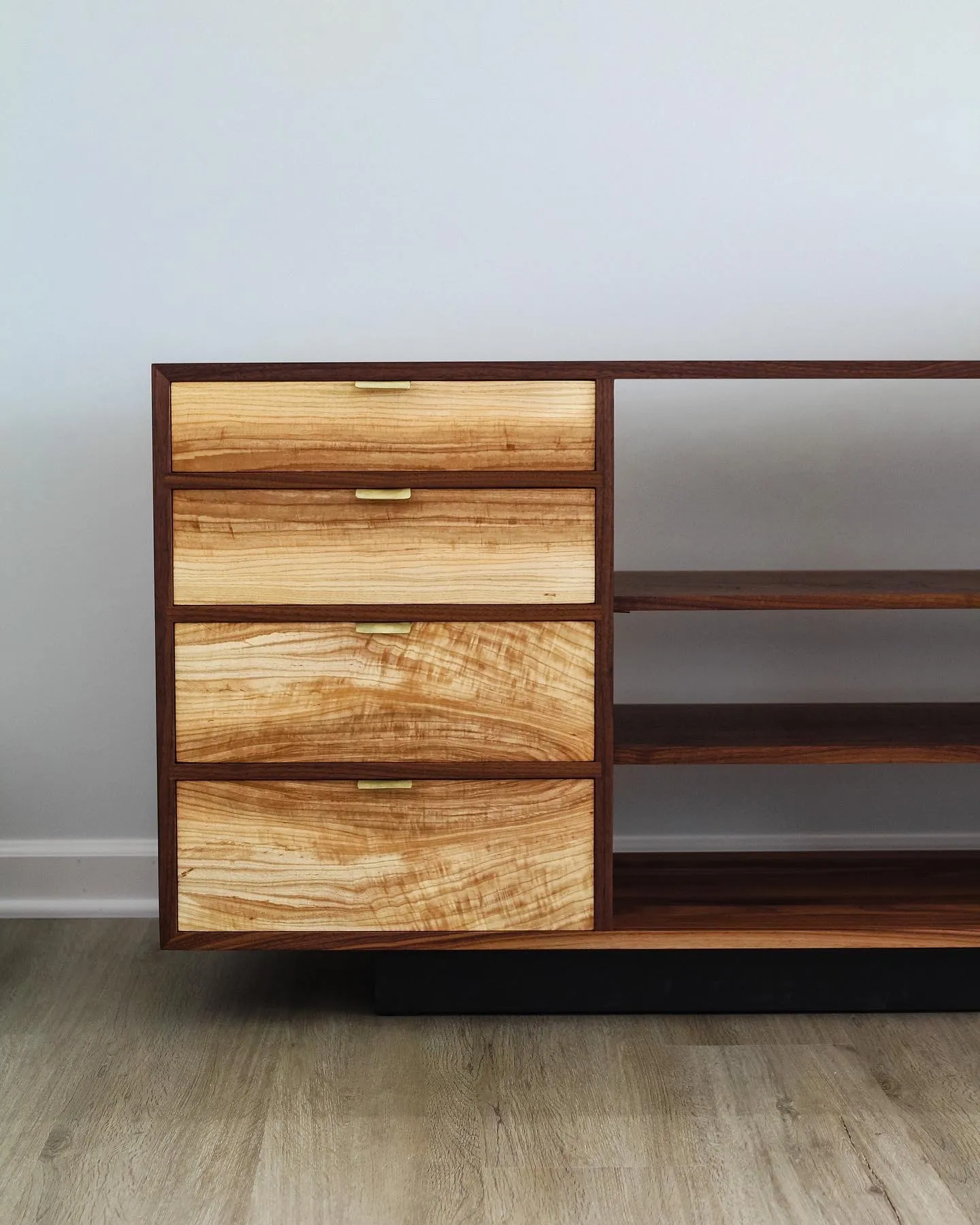 Four ambrosia maple drawer fronts with slim brass pulls, set into a walnut case beside two open shelves.