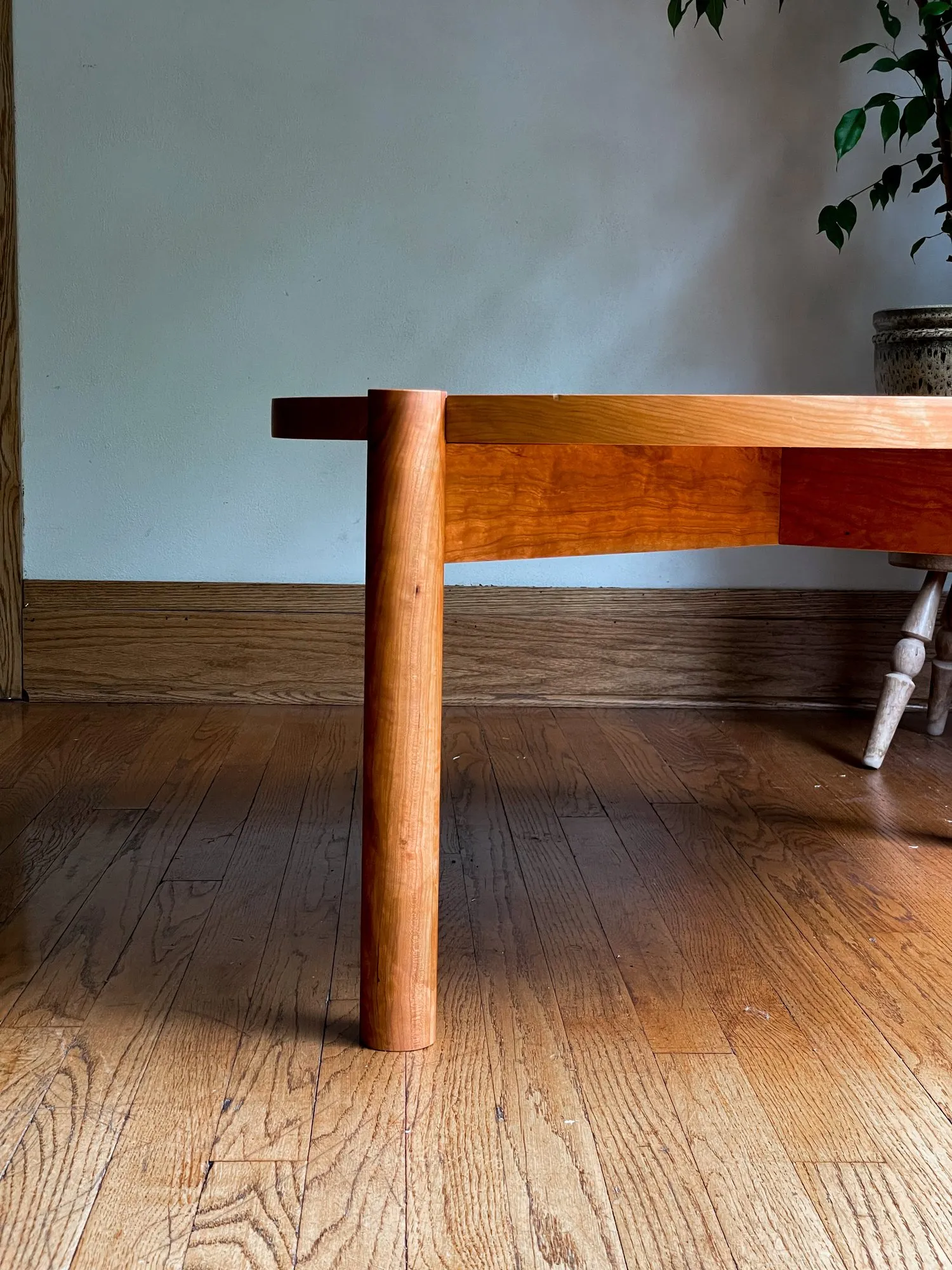 Side profile of the coffee table against a pale wall, showing the thin round top and one slightly tapered turned leg reaching to the floor.