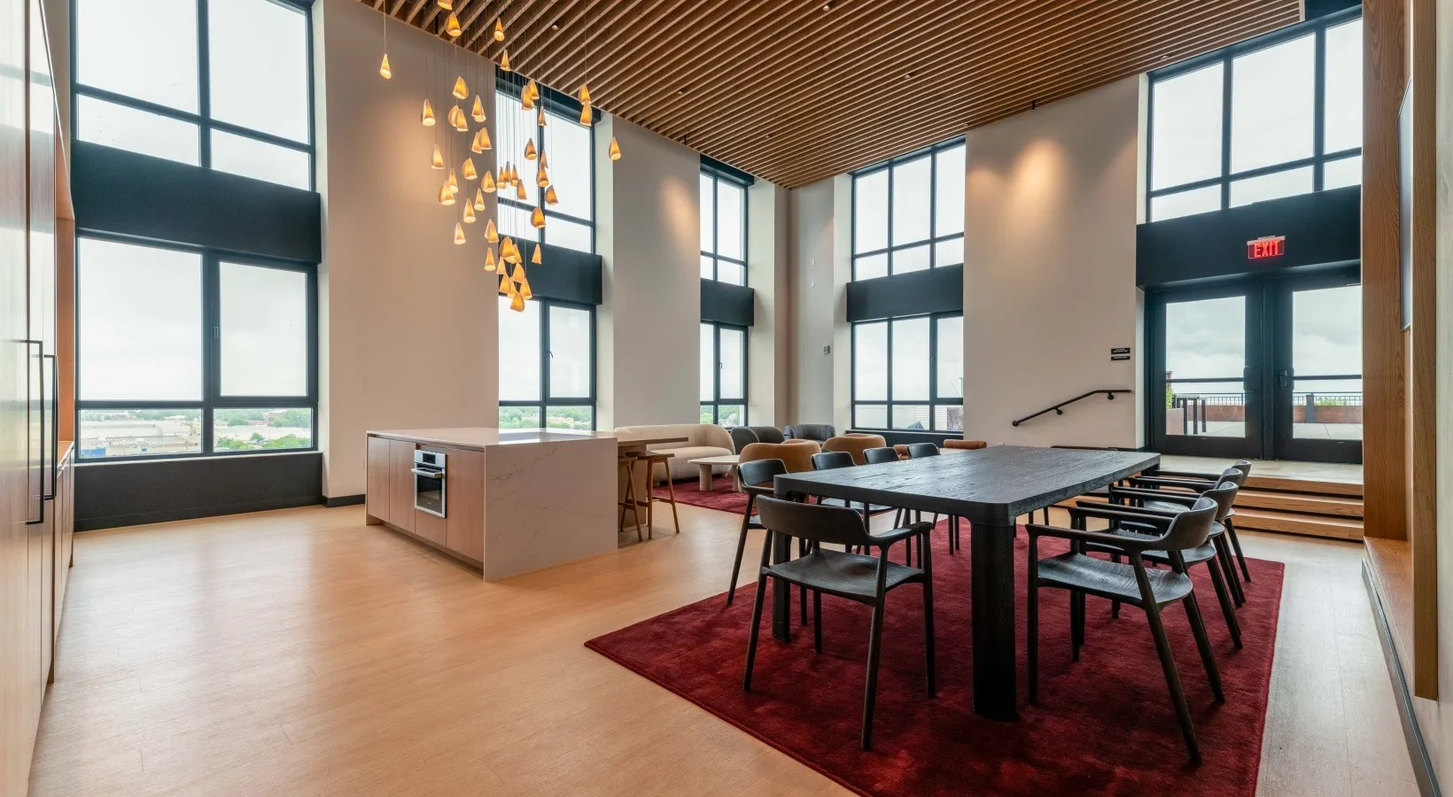 A double-height community room with a wood-slat ceiling, tall windows, a cluster of pendant lights, a stone island, and a long dining table.