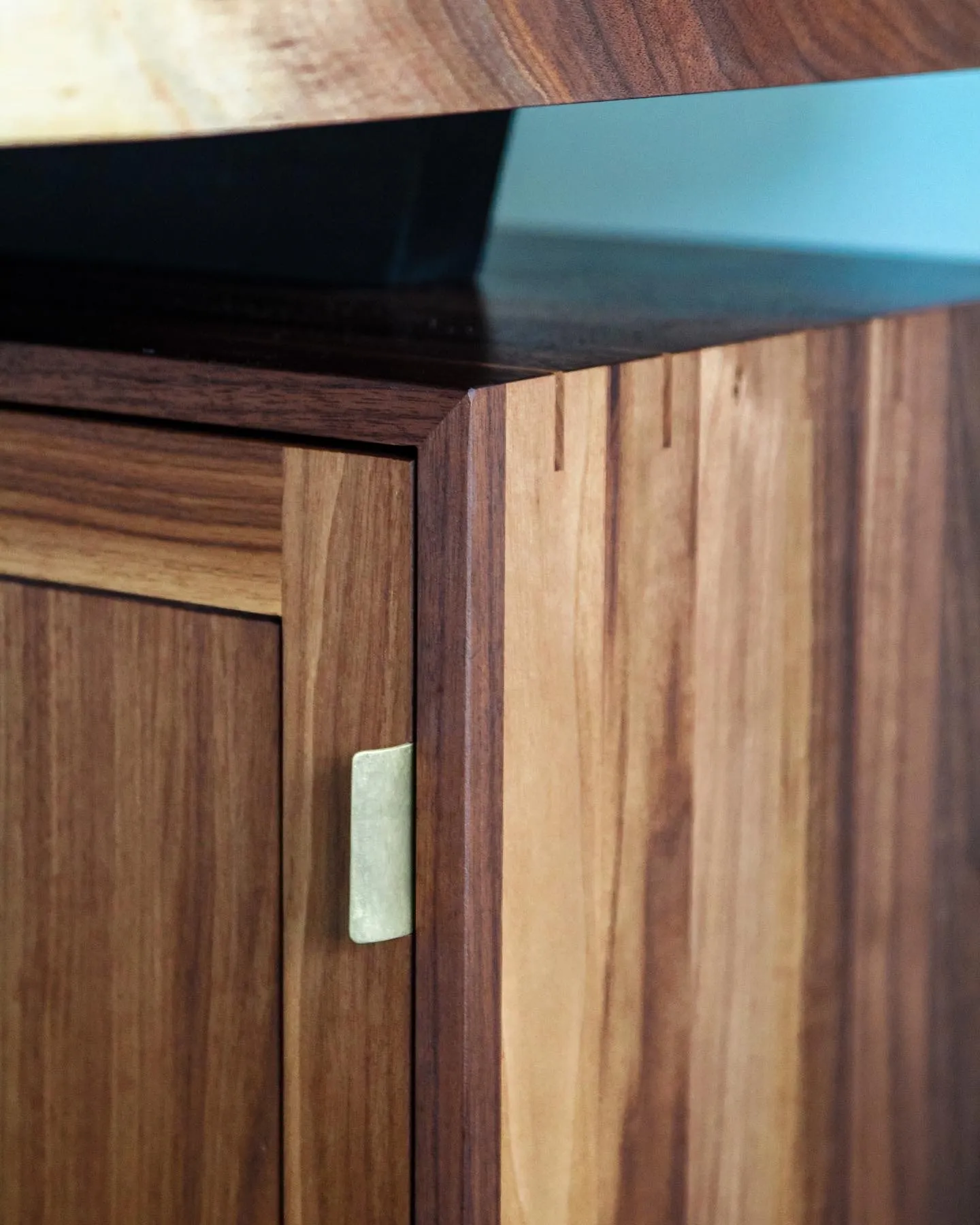 A cabinet door corner with a hand-shaped brass pull, showing the mitered walnut frame and keyed corner joinery.