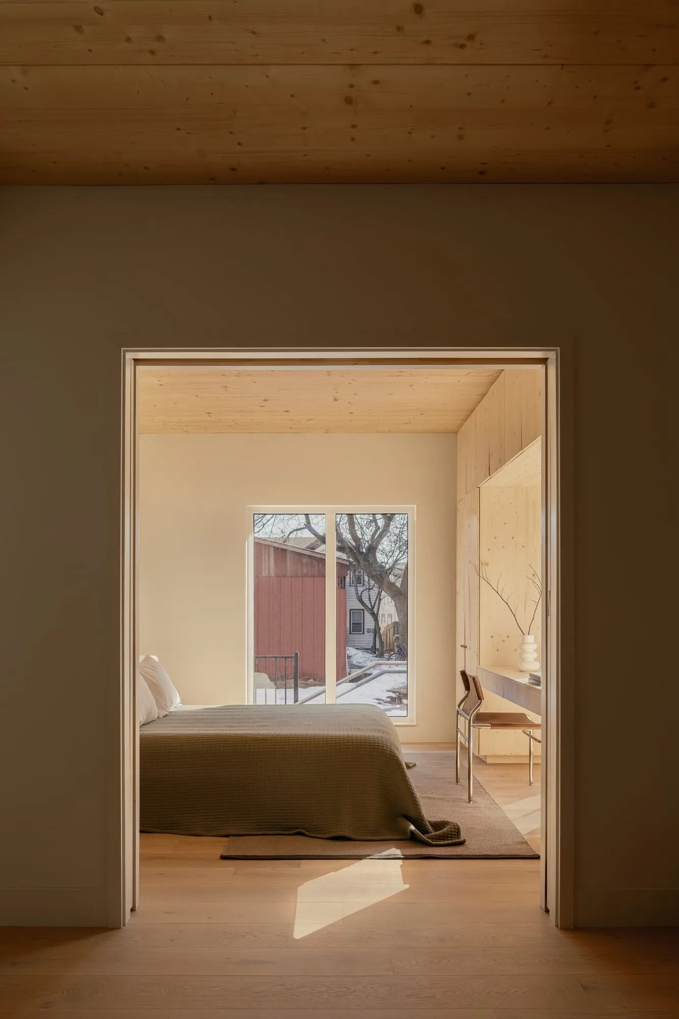 The pine-lined bedroom seen through a doorway, with the wardrobe run and desk nook to one side and a window onto a snowy yard.