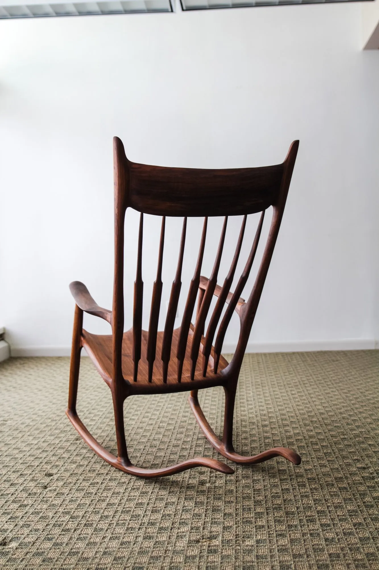 The rocking chair seen from behind, showing the curved crest rail, the fan of spindles, the shaped arms, and the runners sweeping out front and back.
