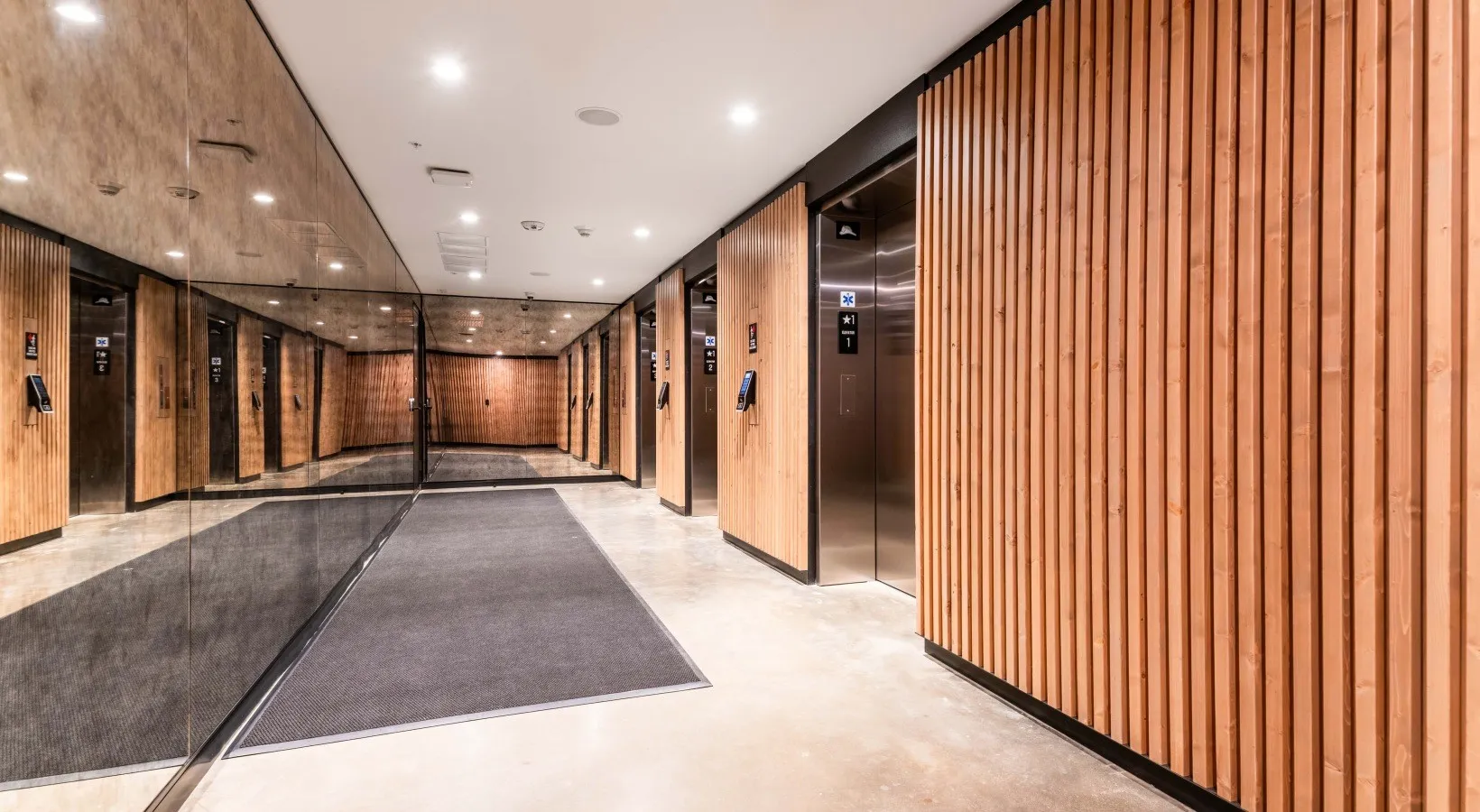 An elevator lobby wrapped in vertical wood slats, a mirrored wall reflecting the run of cab doors.