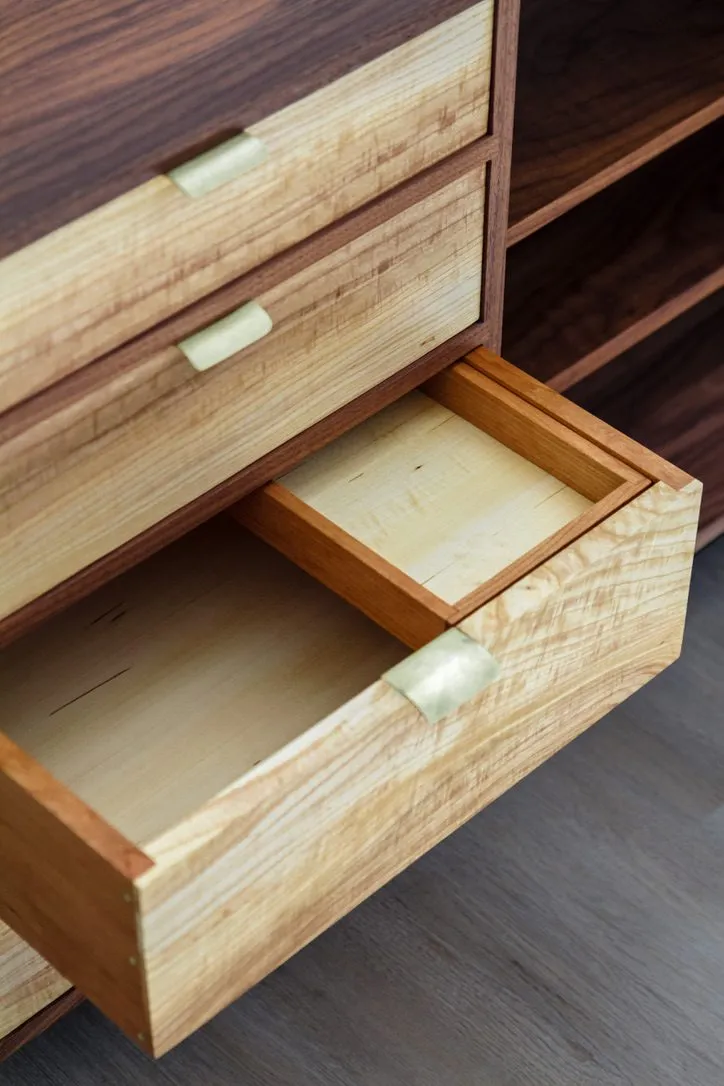 An open drawer showing an ambrosia maple front, a hand-shaped brass pull, and a fitted tray inside the walnut case.