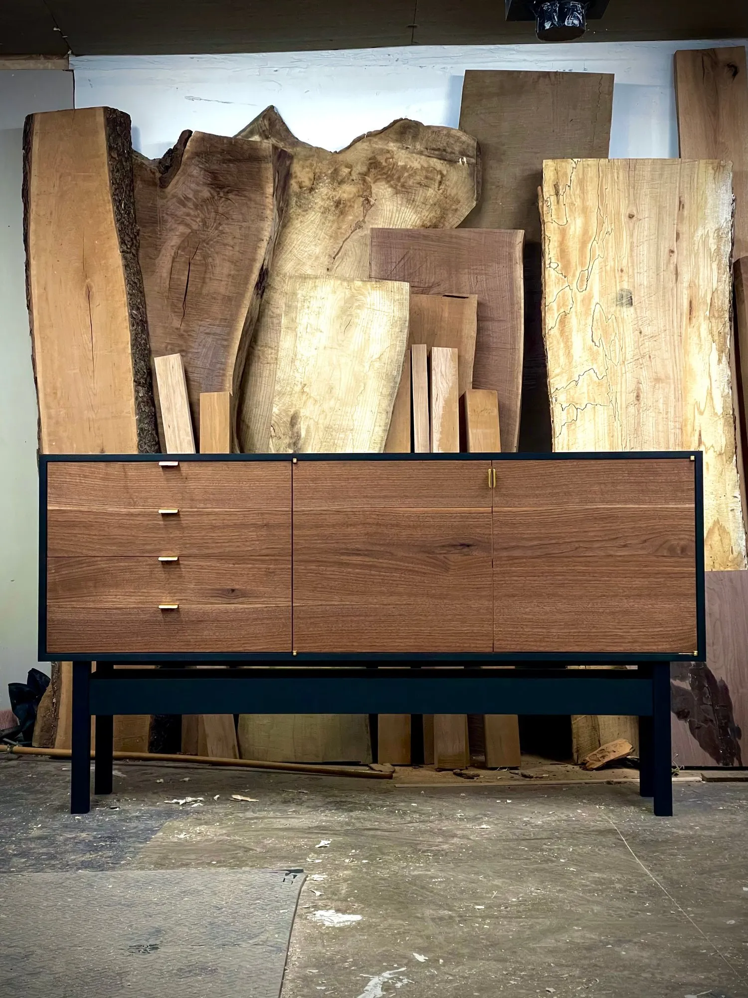 The finished credenza in the Pipermade shop, standing in front of the rough walnut and maple boards it was built from.